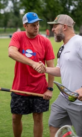 Patrick Chapla in a red POWLAX shirt shaking hands with another coach on a practice field, a lacrosse stick in his other hand.