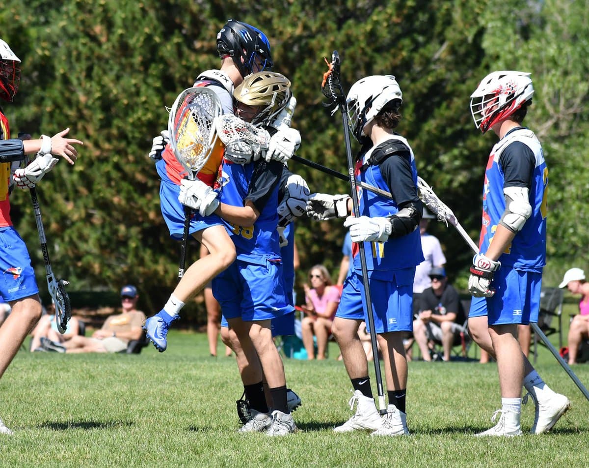 Youth lacrosse players celebrating a goal on the field, two of them embracing while teammates come in around them.